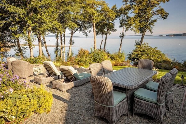 An outdoor seating area with wicker furniture and a table, surrounded by greenery and overlooking a body of water.