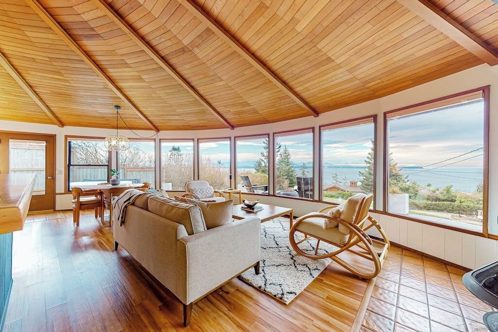 A spacious living room with a wooden ceiling, large windows offering a view of the outdoors, a sofa, a rocking chair, and a dining area.