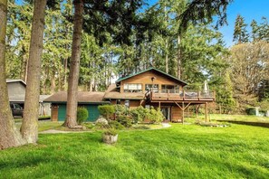 A two-story wooden cabin with a balcony, surrounded by a well-maintained lawn and tall trees. - Scenic Log Cabin with Hot Tub, Beach Access and Game Room (Freeland)