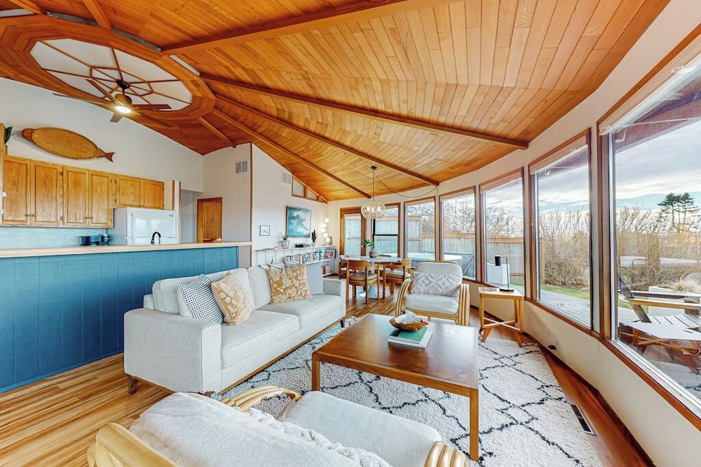 Amplia sala de estar con techo de madera, cocina con armarios azules y ventanas grandes con vistas al exterior.
