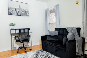 A room with a black sofa, a desk with a chair, a cityscape picture on the wall, and a window with curtains. - Cozy 2-bedroom family friendly duplex in quaint East Hartford with office space (East Hartford)