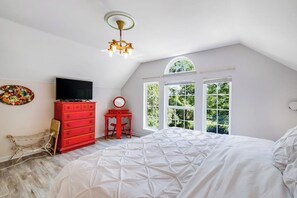 Ein Schlafzimmer mit schrägen Wänden, einem großen Bett, einer roten Kommode, einem Fernseher und einem Fenster mit Blick auf Grünflächen.