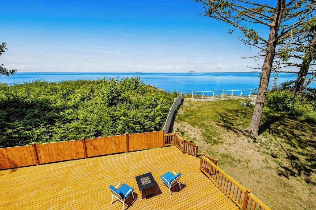 A wooden deck with two chairs and a table, overlooking a coastal landscape with a clear view of the sea and distant land.
