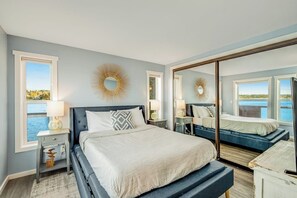 A bedroom with a large bed, bedside tables, a mirror, and a view of the outdoors. - Waterfront Family Friendly Home with Bay Views, Firepit, Deck, & Fireplace (Coupeville)