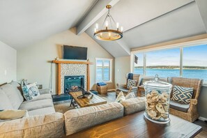 Un salon chaleureux avec une cheminée, une grande fenêtre donnant sur un plan d’eau et un lustre.