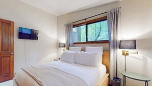 Een hotelkamer met een bed, een aan de muur bevestigde televisie, een raam met gordijnen, een bedlamp en een klein tafeltje.