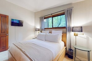 A hotel room with a bed, a television mounted on the wall, a window with curtains, a bedside lamp, and a small table. - Pet Friendly Mountain View Retreat on Orcas Island with BBQ and Patio (Orcas Island)