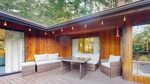 Een houten terras met rieten meubels en een glazen tafel, omgeven door grote ramen.