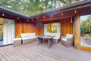 A wooden deck with wicker furniture and a glass table, surrounded by large windows. - Pet Friendly Mountain View Retreat on Orcas Island with BBQ and Patio (Orcas Island)