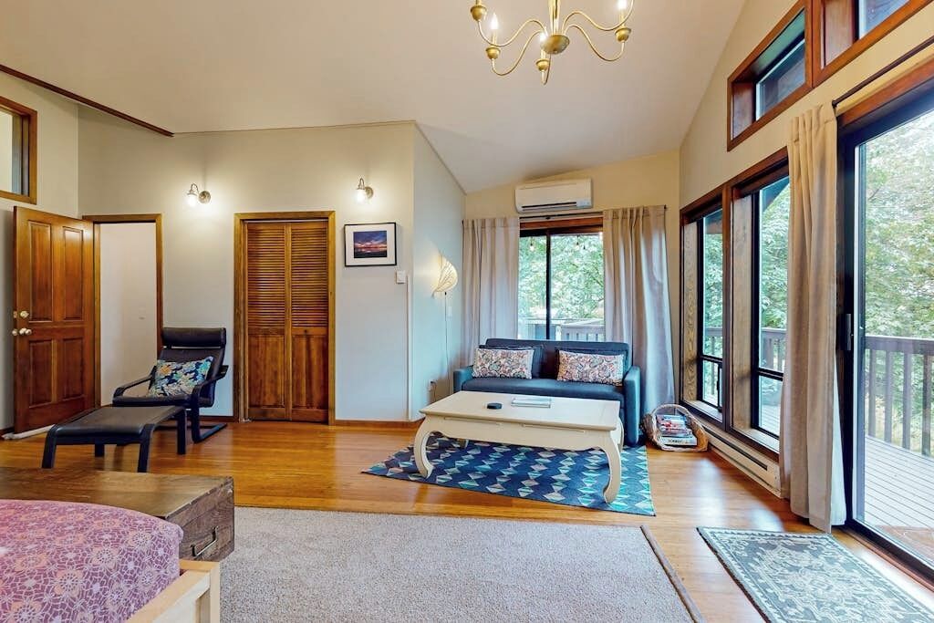 A spacious room with a large bed, a sofa, and a dining table. There are multiple windows providing natural light and a view of the outdoors.