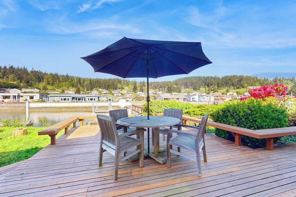 Eine Holzterrasse mit einem runden Tisch, Stühlen, einem Sonnenschirm und Bänken.