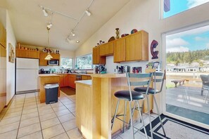 A kitchen with wooden cabinets, a central island, bar stools, and a large window offering a view. - Peaceful Island Retreat with Community Dock on Whidbey Island (Oak Harbor)