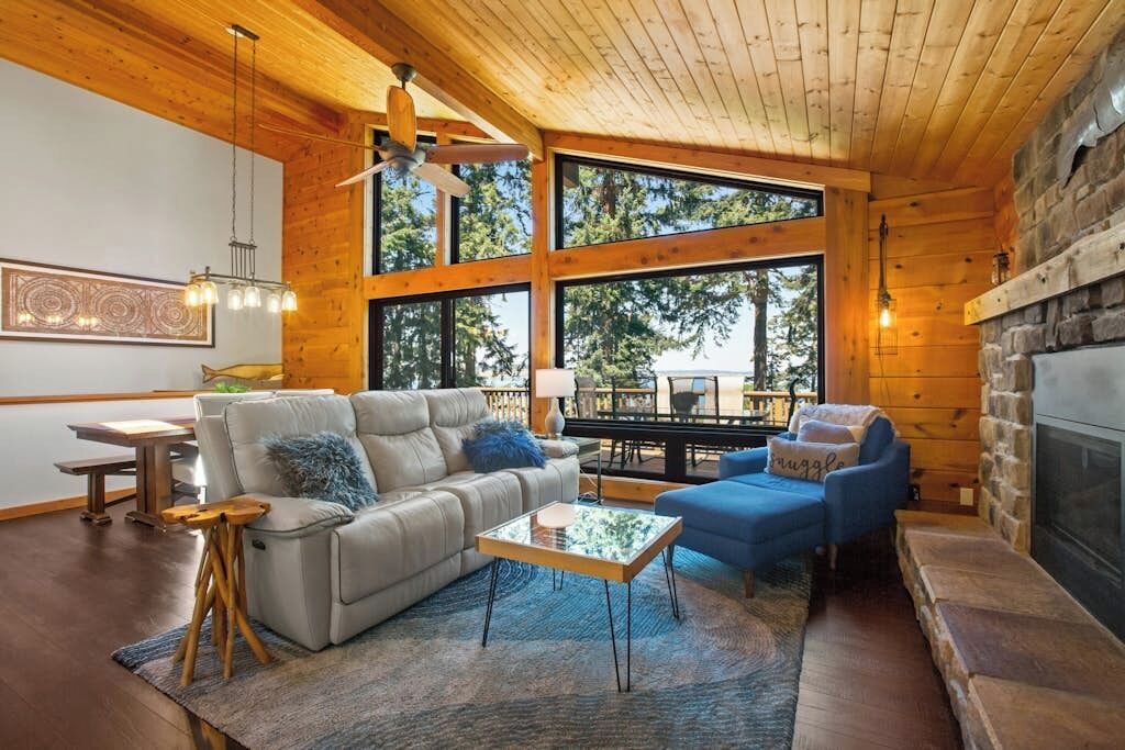A cozy living room with a fireplace, large windows, and a ceiling fan.