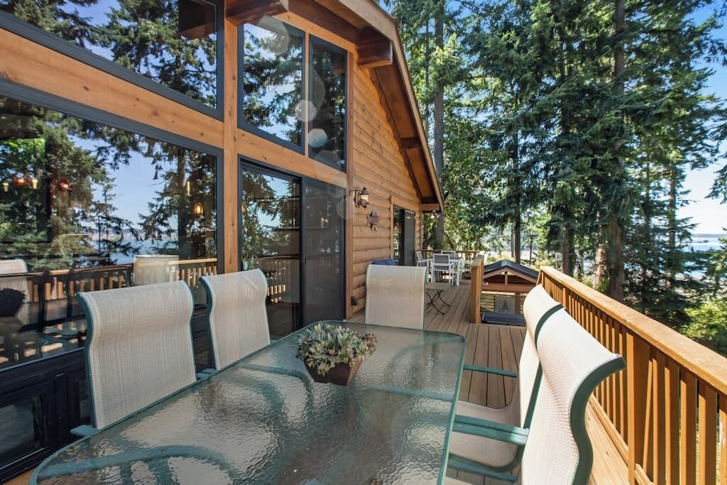 A wooden deck with a glass table and chairs, surrounded by trees.