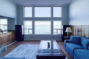 A room with a large window overlooking the sea, a blue sofa, a glass coffee table, and a wooden cabinet.