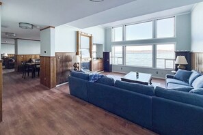 A spacious living area with a large window overlooking a body of water, a blue sofa, a dining table with chairs, and a kitchen area.