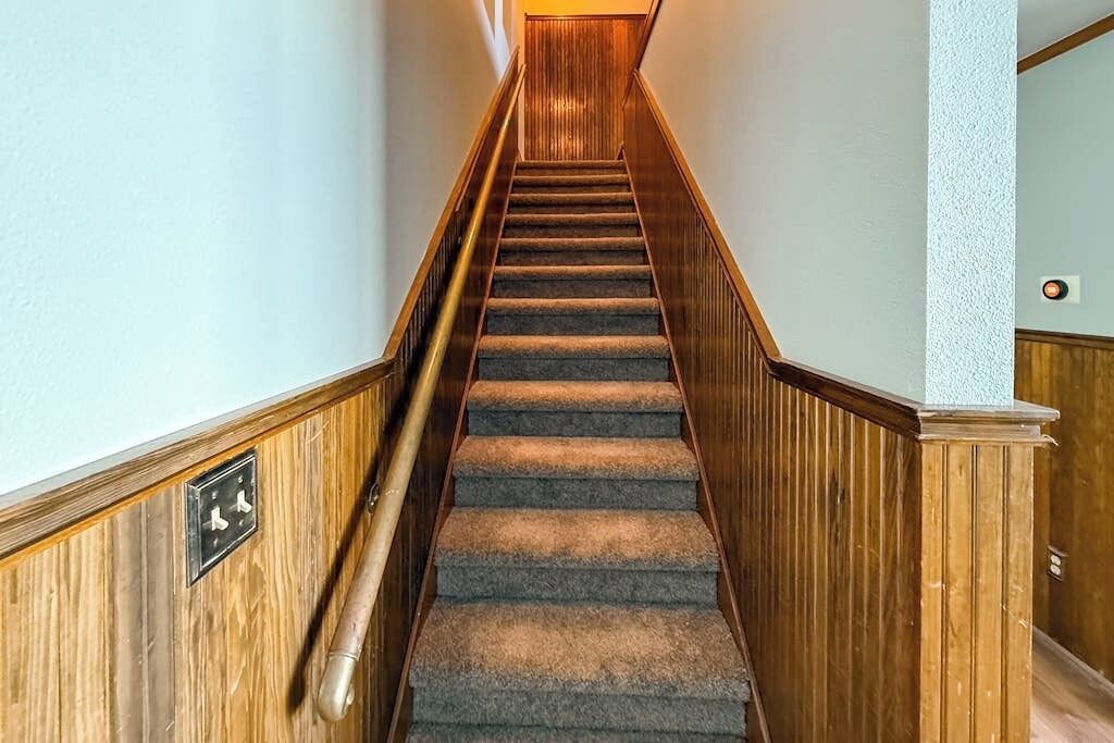 Un escalier recouvert de moquette, avec des rampes en bois et un interrupteur sur le mur.