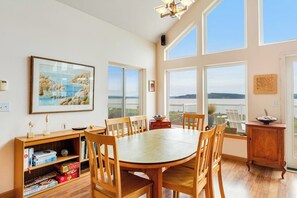 A dining area with a round table, wooden chairs, a bookshelf with board games, and large windows offering a view. - Stunning 4 Bedroom Beachfront Home in Greenbank with Panoramic Water Views (Greenbank)