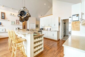 A modern kitchen with white cabinets, a central island with bar stools, and hanging pots. - Stunning 4 Bedroom Beachfront Home in Greenbank with Panoramic Water Views (Greenbank)