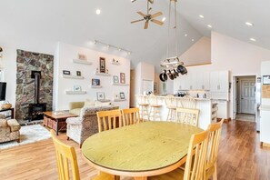 A spacious living area with a stone fireplace, a dining table with chairs, and a kitchen with white cabinets and a hanging pot rack. - Stunning 4 Bedroom Beachfront Home in Greenbank with Panoramic Water Views (Greenbank)