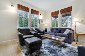 Ein Wohnzimmer mit einer Couch, zwei Sesseln, einem Couchtisch und einem Teppich. Es gibt Fenster mit Jalousien und eine Lampe auf einem Beistelltisch.