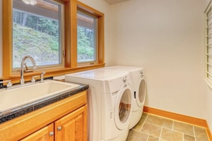A laundry room with a sink, two washing machines, wooden cabinets, and a window with a view of trees.