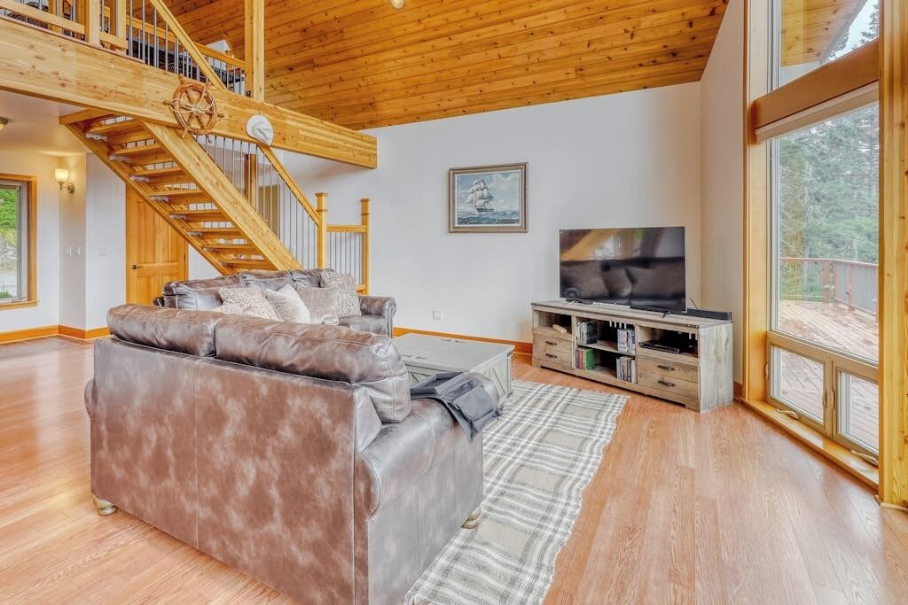 A spacious living room with a large leather sofa, a wooden staircase, a flat-screen TV, and a large window.