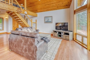 A spacious living room with a large leather sofa, a wooden staircase, a flat-screen TV, and a large window.