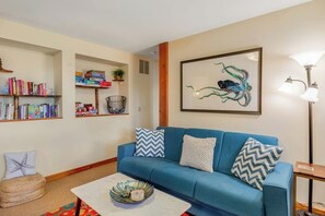 A living room with a blue sofa, a coffee table, a bookshelf, and a framed artwork. - Scenic Waterfront Home with Hot Tub and Three Unique Living Spaces (Coupeville)