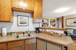 Cocina compacta con armarios de madera, un lavamanos y una encimera. Se observa una cafetera, un hervidor y una pequeña estantería con libros y adornos.