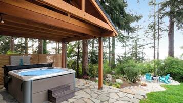 A hot tub under a wooden pergola with stone flooring, surrounded by greenery and outdoor furniture.