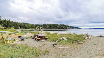 A beach with a picnic table and chairs, a boat, and a forested hill in the background.