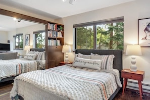 A bedroom with two beds, a bookshelf, a TV, and a large window with a view of trees.
