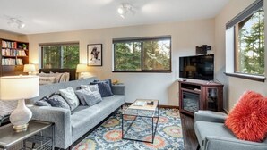 Una sala de estar con sofá, mesa de centro, televisor, estantería y ventanas amplias.