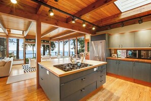 A modern kitchen with a central island, wooden ceiling beams, and large windows overlooking a scenic view. - Exquisite Oceanfront Home with Balcony, and Gym in Freeland (Freeland)