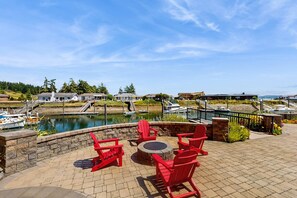A waterfront patio with red Adirondack chairs, a stone fire pit, and a view of boats and houses across the water. - Stunning Waterfront Home with Private Dock and Sauna in Oak Harbor (Oak Harbor)