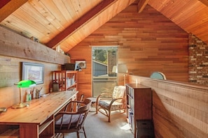 Ein gemütliches Dachzimmer mit Holzbalkendecke, einem Schreibtisch mit Stühlen, einem Bücherregal und einem Fenster.