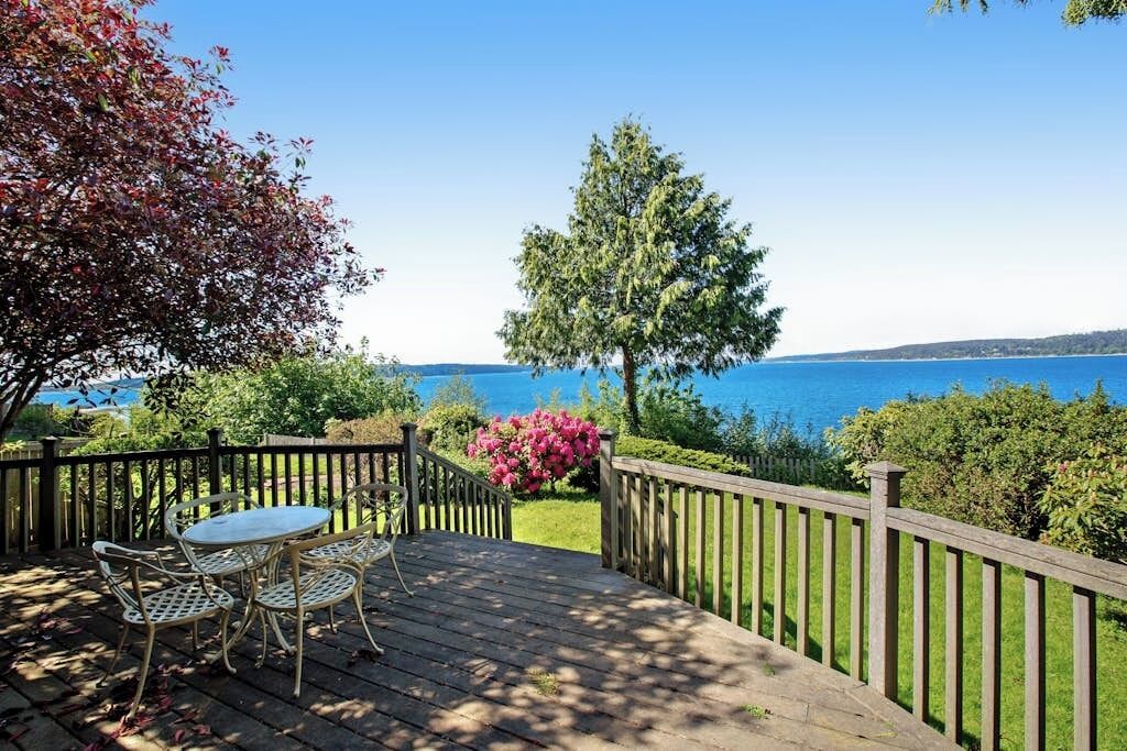 Un terrazzo in legno con tavolo e sedie, affacciato su uno specchio d'acqua e un cielo sereno.