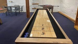 A shuffleboard table with numbered scoring areas, a foosball table, and a table with chairs in a room with white walls and a blue carpet. - 4 BR Pet Friendly Waterfront Home with Game Room (Clinton)