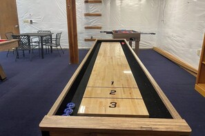 A shuffleboard table with numbered scoring areas, a foosball table, and a table with chairs in a room with white walls and a blue carpet.