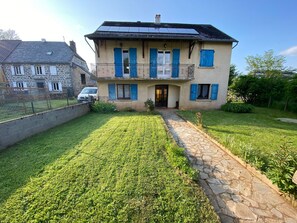 A two-story house with a stone pathway leading to the entrance, surrounded by a well-maintained lawn and a fenced garden area. - Country Cottage Near Pierrefort and Col de Prat de Bouc (Cézens)