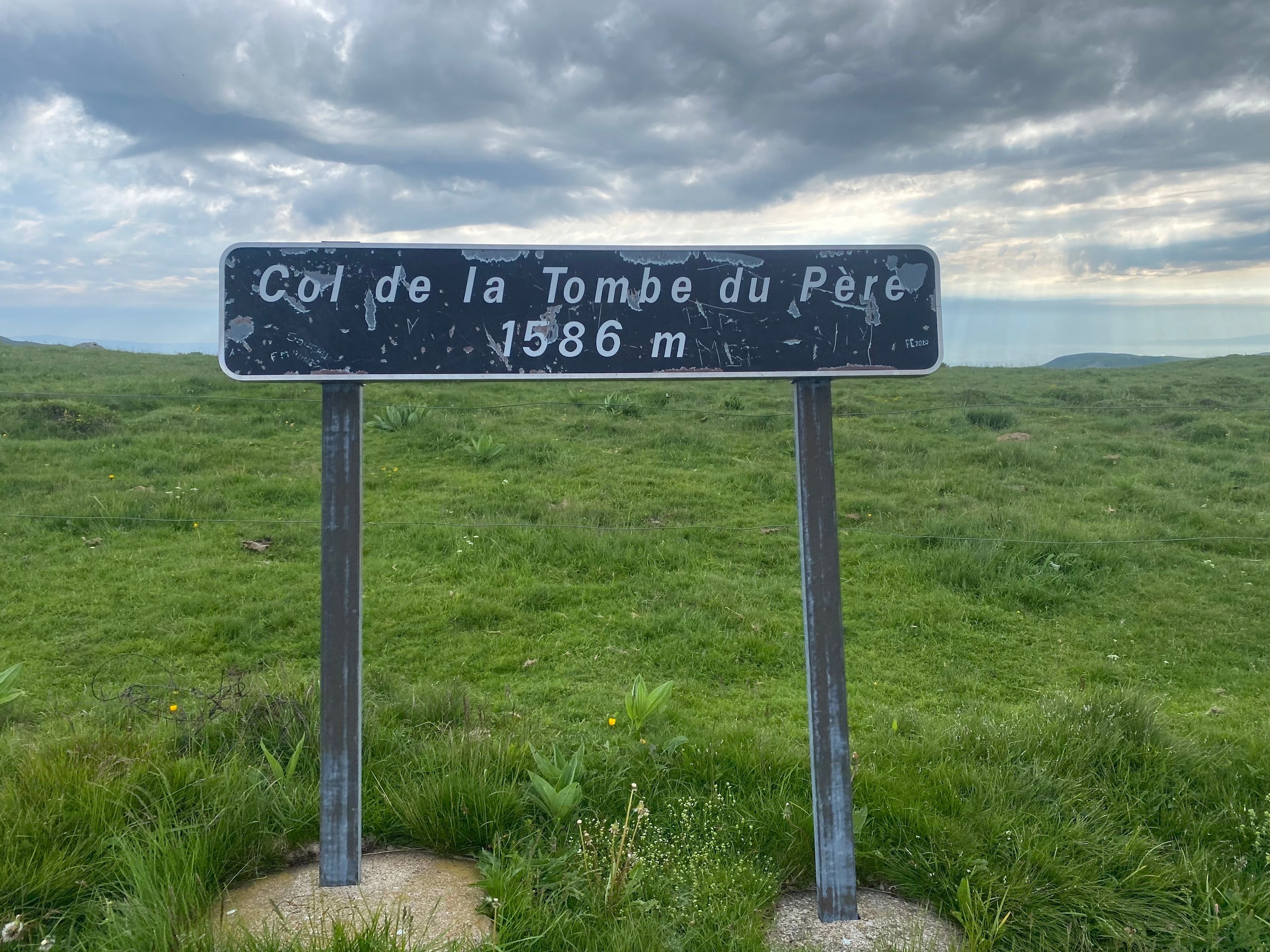 Um sinalizador com a inscrição 'Col de la Tombe du Père 1586 m' em um campo gramado.