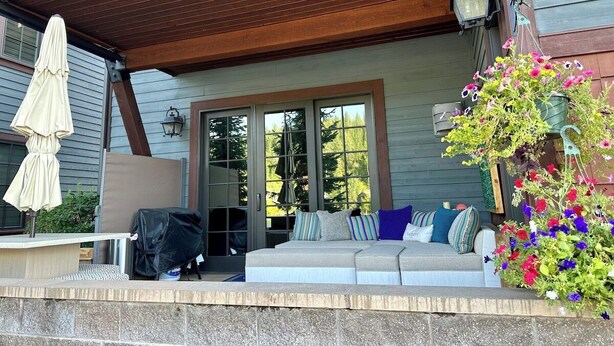 A covered outdoor seating area with a sofa and cushions, a covered table, and a hanging flower planter. - Vesper Bleu  2 Kings + EV Charger (Coeur d'Alene)