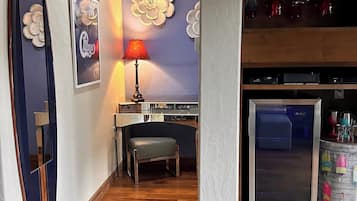 A hotel lobby with a wooden floor, a desk with a chair, a refrigerator, and a wall with decorative elements.