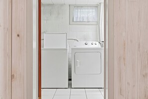 Ein Waschraum mit Waschmaschine und Trockner, weißen Fliesenwänden und einem Fenster mit weißem Vorhang.