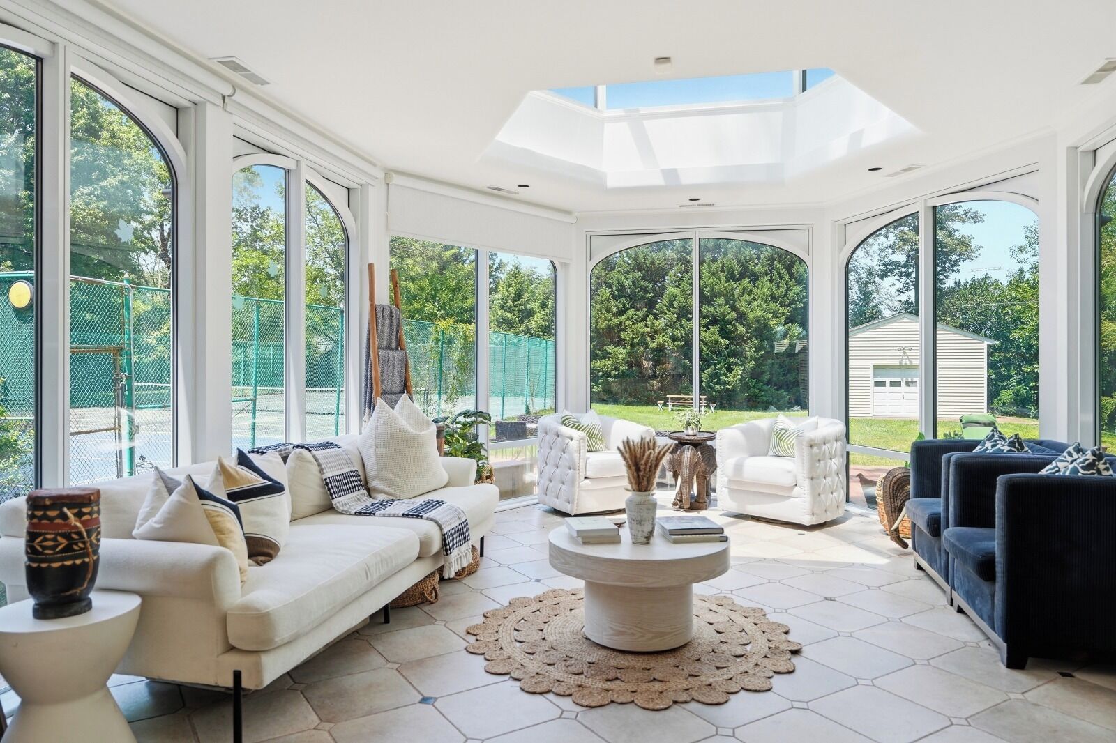 A spacious sunroom with large windows, a white sofa, a blue sofa, and a round coffee table.
