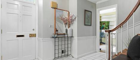 A hallway with a white door, a console table with a mirror and vase, a staircase with a wooden handrail, and a tiled floor.