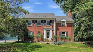 A two-story brick house with a front porch, a garage, and a well-maintained lawn. - 
The Bishop Retreat  5BR, Tennis Court & Yard (Alexandria)