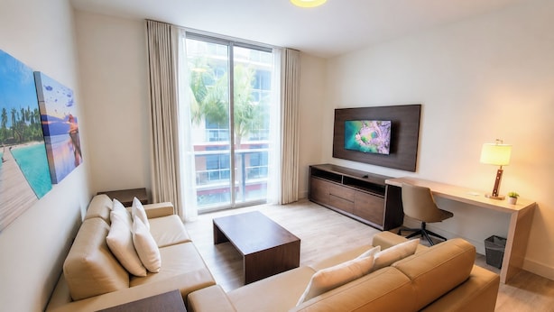 Ein modernes Wohnzimmer mit einer Sofagarnitur, einem Couchtisch, einem an der Wand montierten Fernseher und einem Schreibtisch mit einer Lampe.