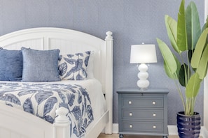 A bedroom with a white bed, blue patterned bedding, a bedside table with a lamp, and a potted plant. - New Listing! Updated Oceanfront 2/2 | Heated Pool & Garage Parking (Ormond Beach)
